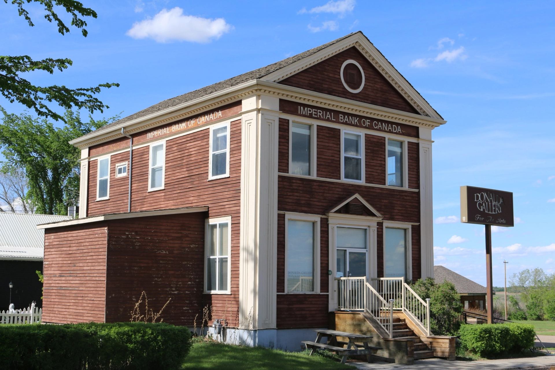A heritage building with a sign reading "Donalda Gallery For The Arts" located on a street corner. The building has large windows, a staircase, and is surrounded by trees and green landscaping.