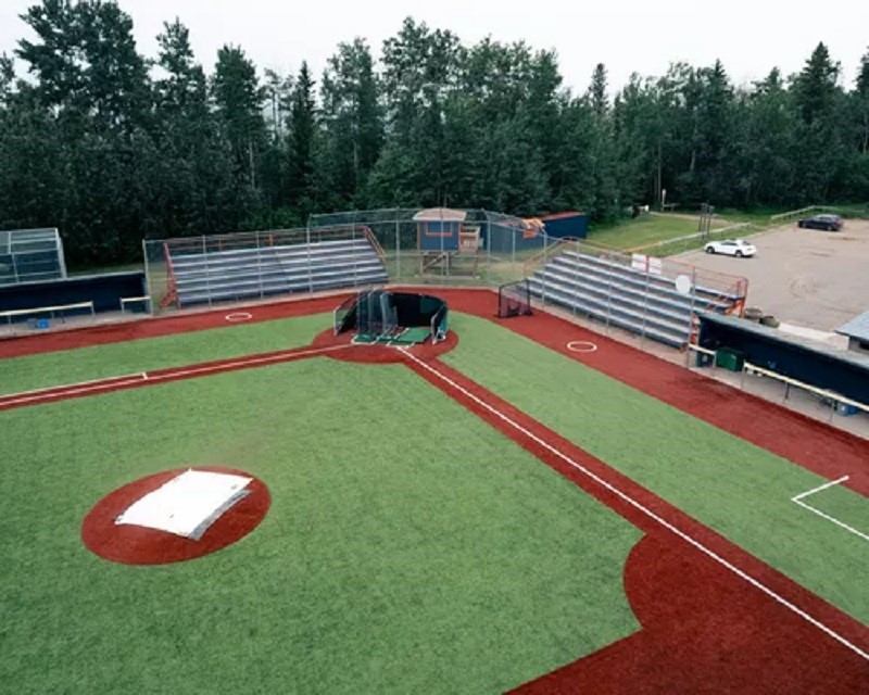Baseball diamond with artificial turf, bleachers, backstop, trees and parking lot.
