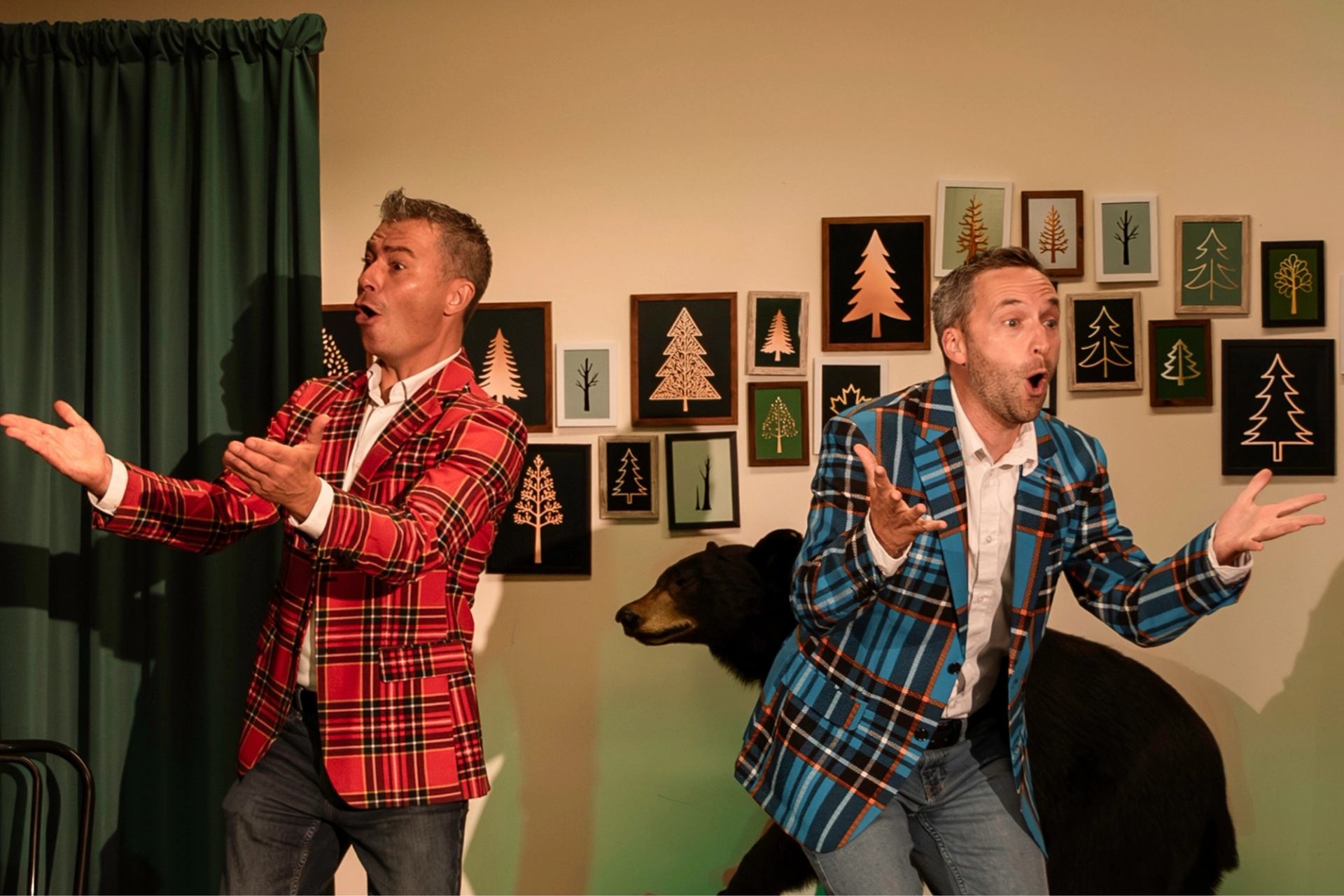 Two performers in plaid jackets gesturing onstage with forest‑themed decor
