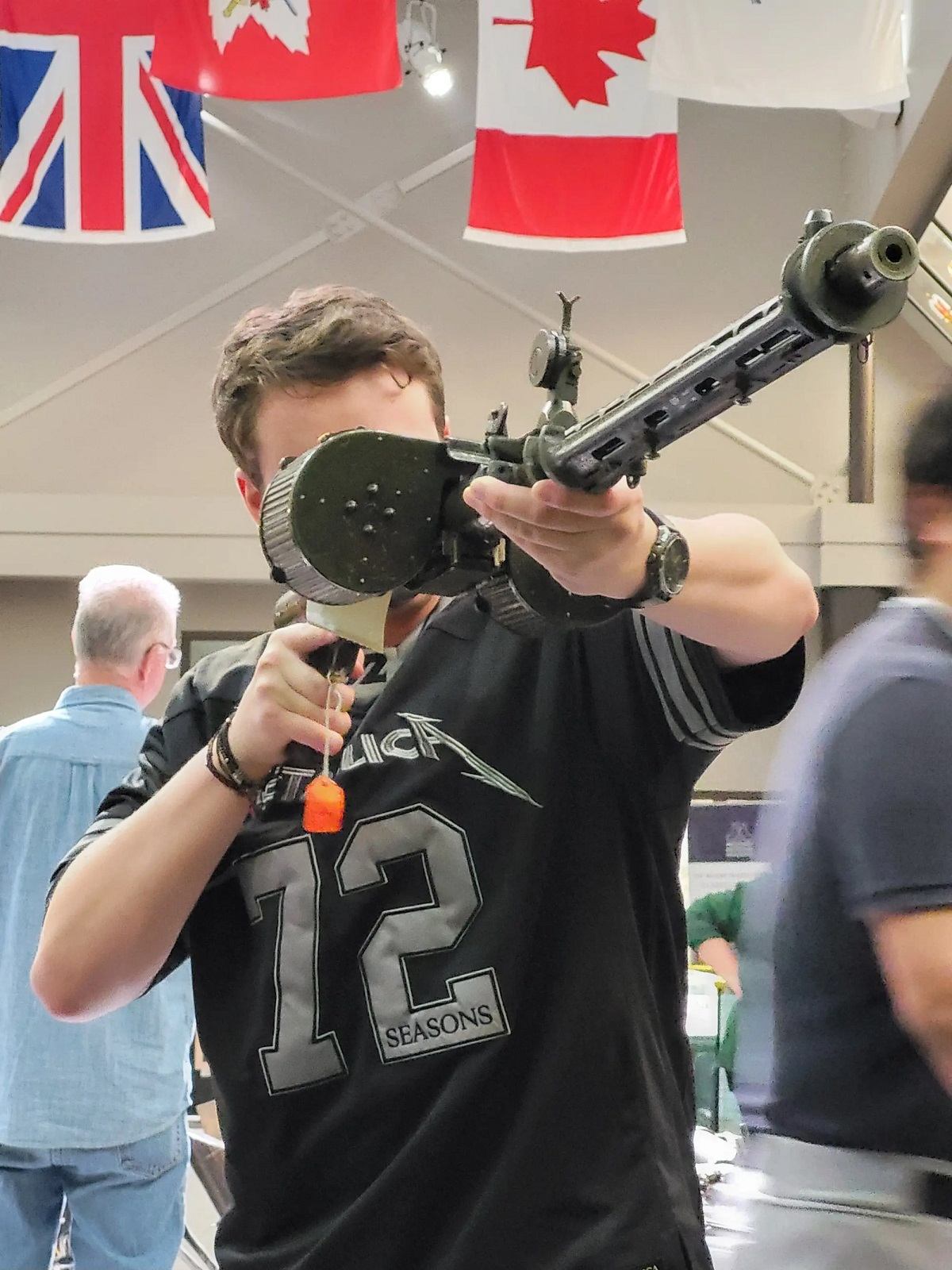 A young man in a black Metallica t-shirt aims a machine gun with a drum magazine. Union Jack and Canadian flags are in the background.