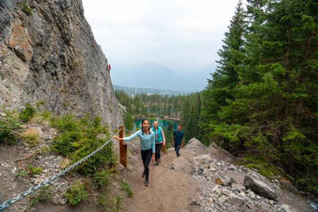 A family hikes up popular local hike Grassi Lakes while a rock climber climbs the rockface directly behind them.