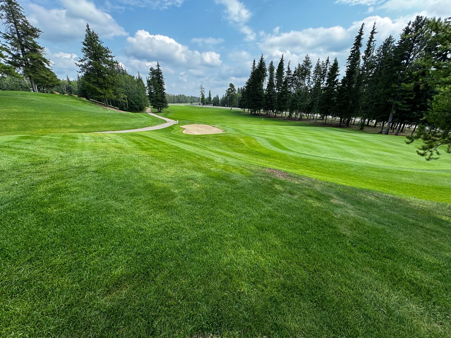 Manicured fairway and green at The Dunes Golf & Winter Club with sand bunker, trees, and blue sky.