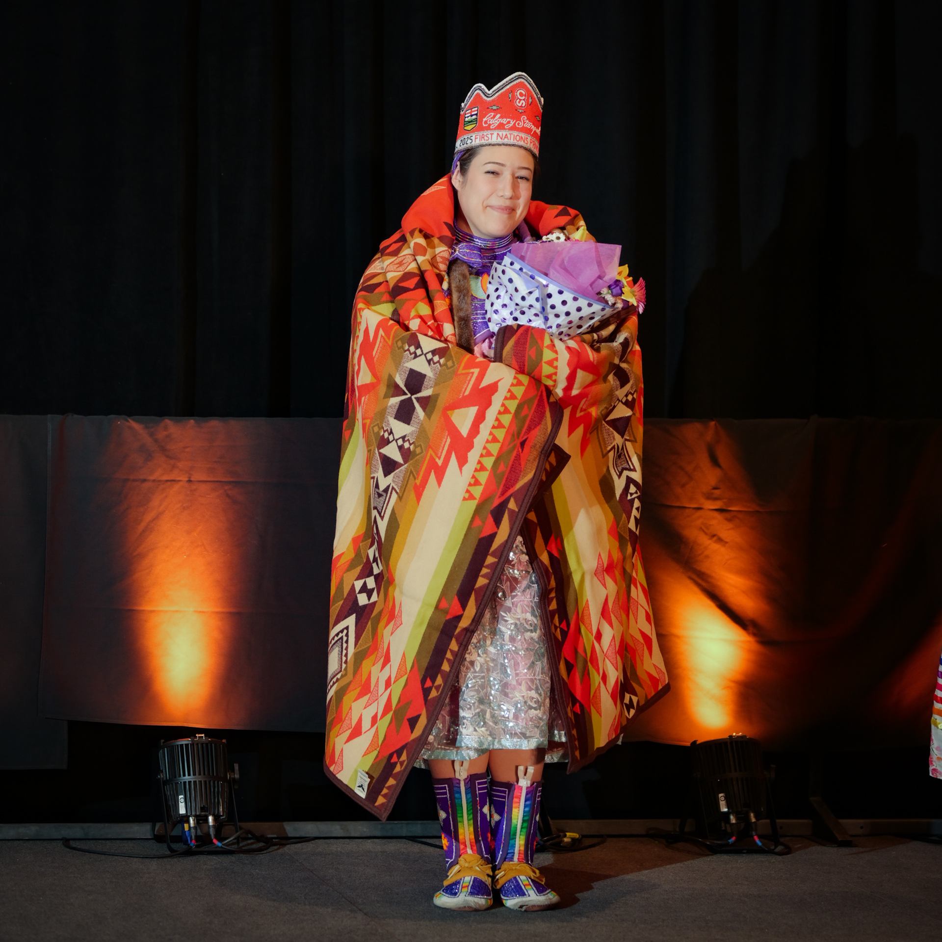 Person in regalia wrapped in patterned blanket holds bouquet on stage with lights.