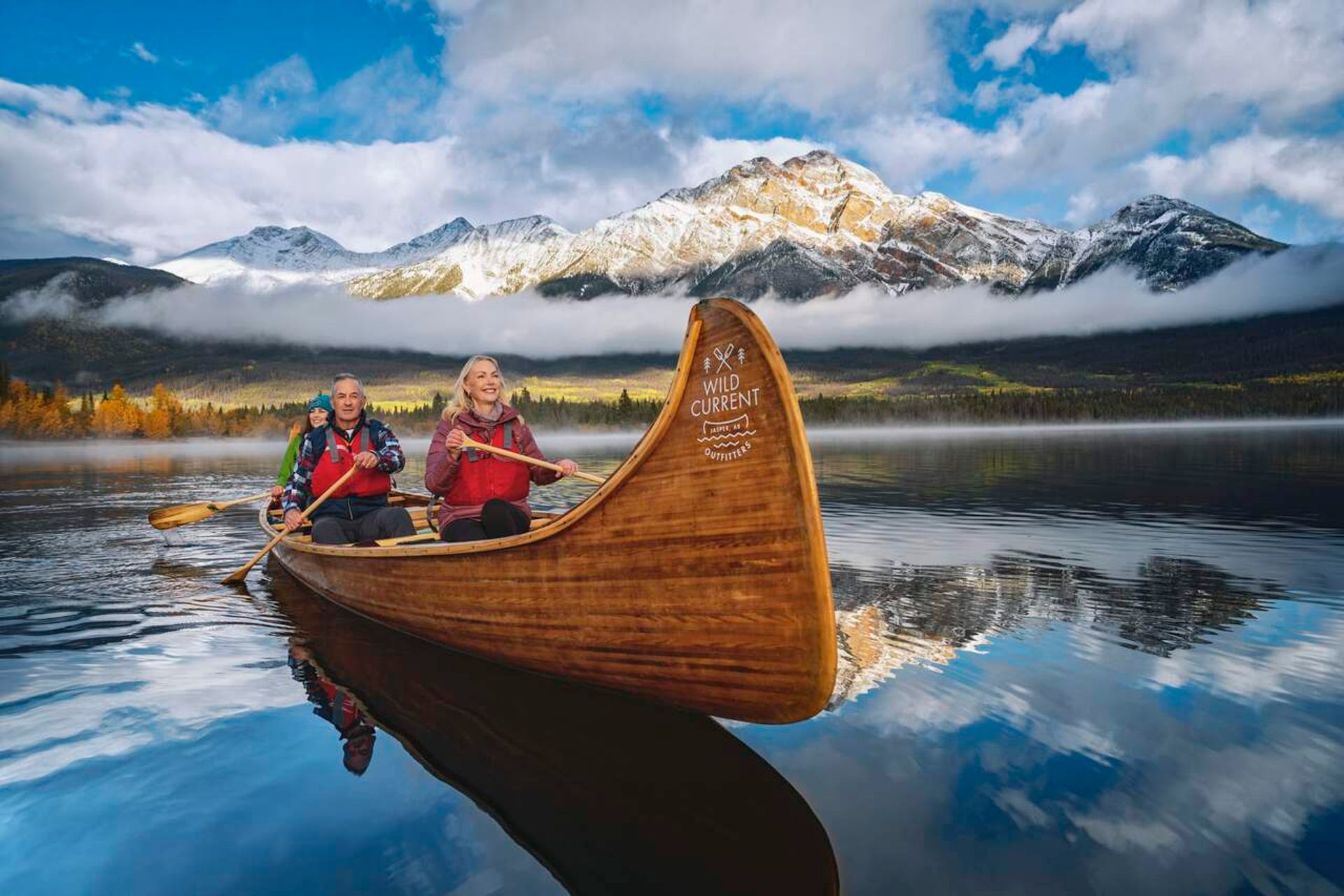 Wooden Wild Current canoe with three paddlers on calm lake, snowy mountains behind.