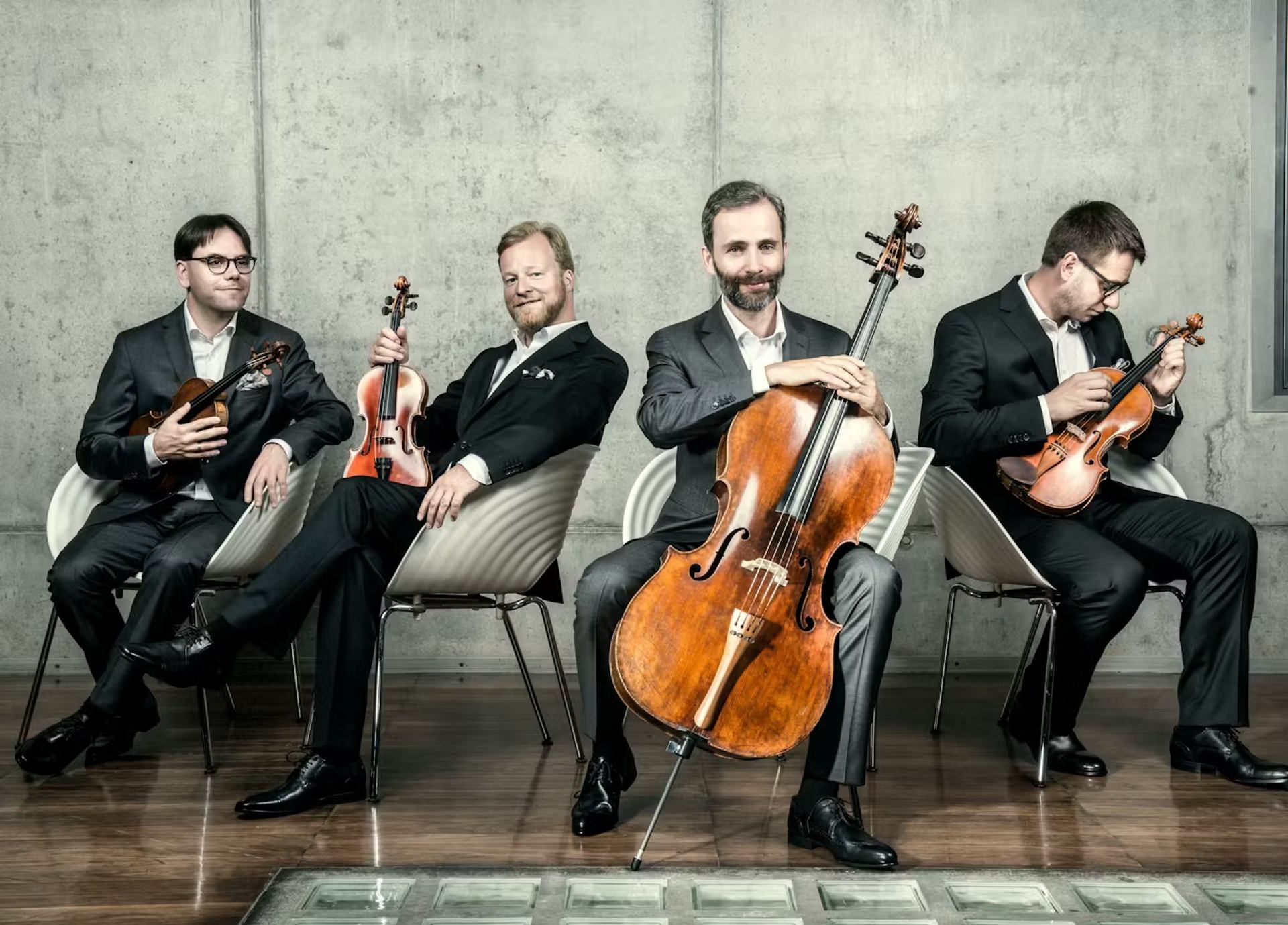 Four string musicians seated with violin, viola, and cello against a concrete wall.
