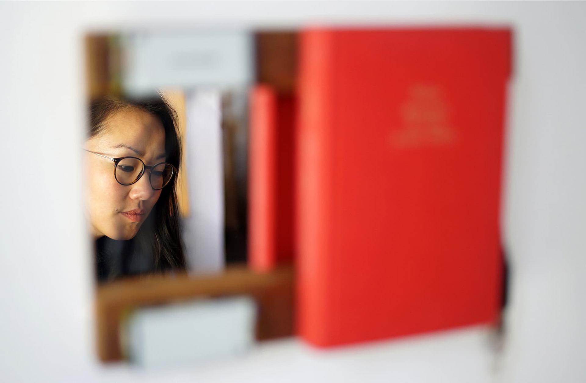 Person wearing glasses reflected in a mirror between blurred red and white books.