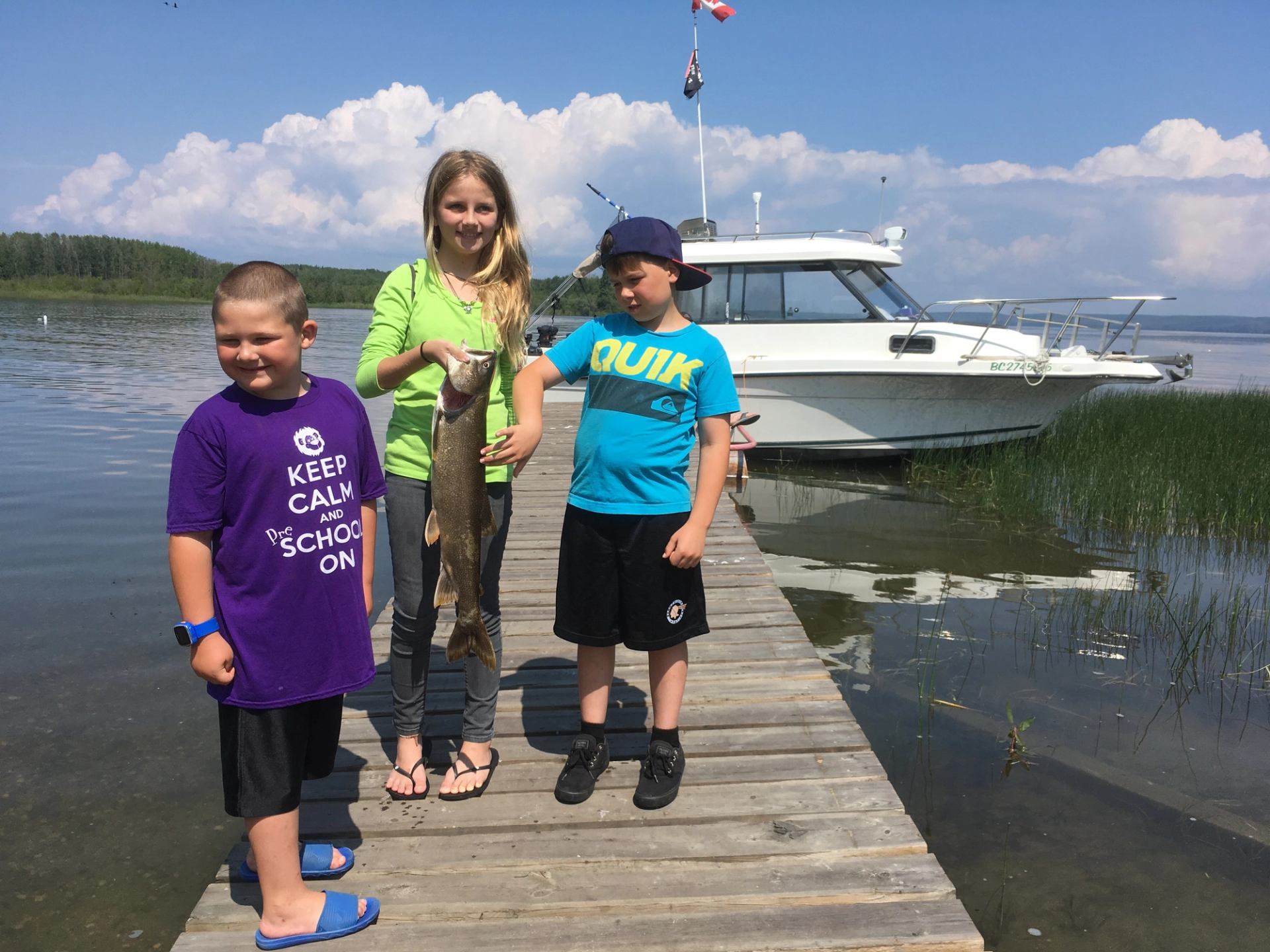 Three kids on a dock, one proudly holding a large fish by a lake.