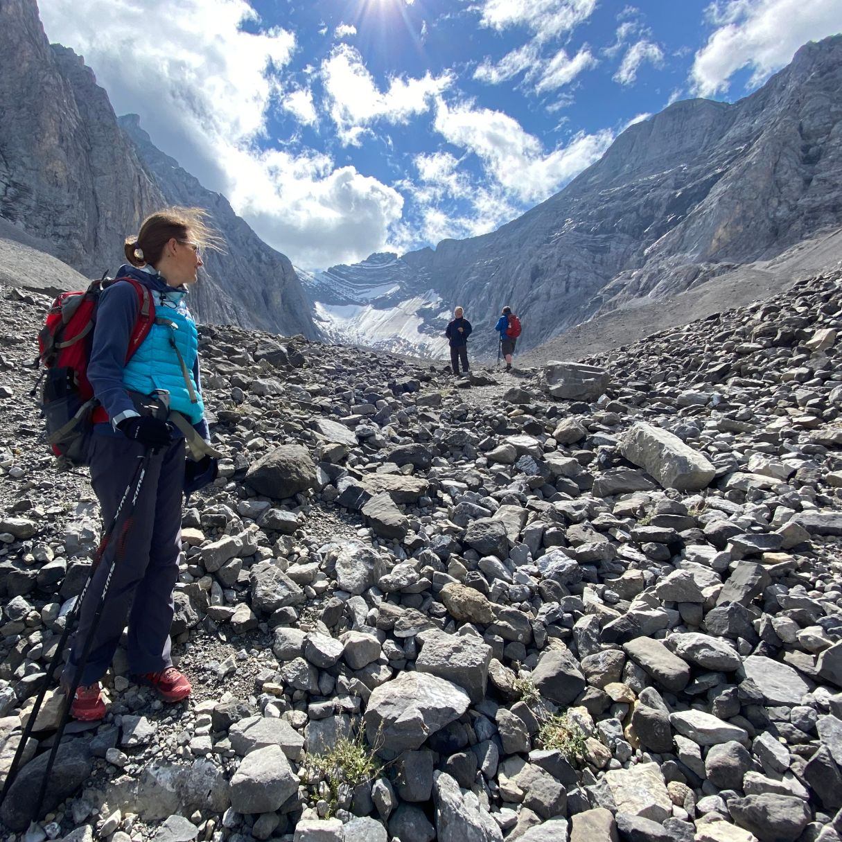 Hikers on rocky mountain trail near a glacier valley under partly cloudy sky.