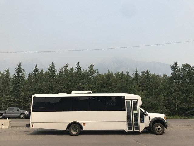 White Jelloun Travel bus with tinted windows and open door, parked near trees and mountains.