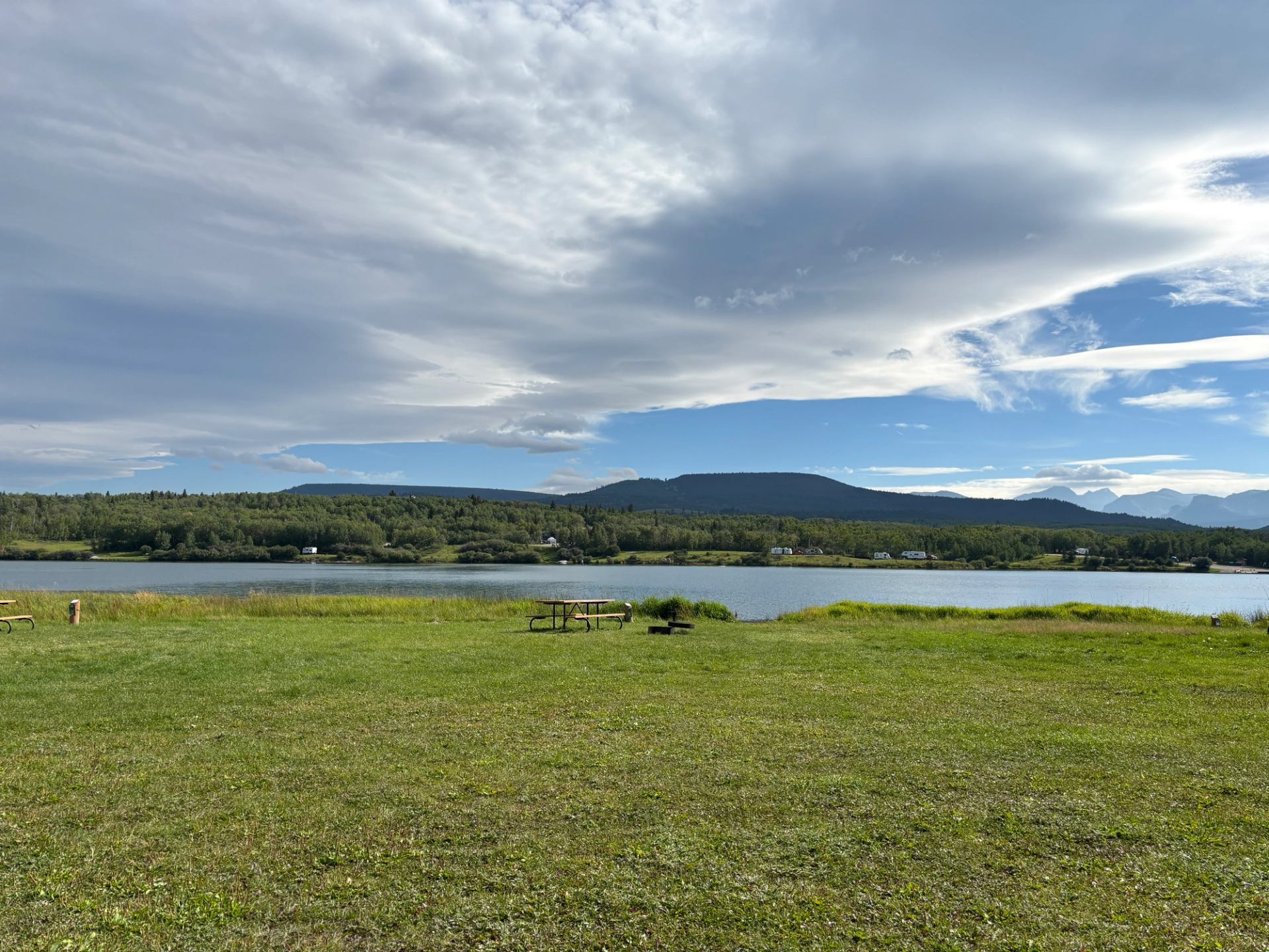 Grassy lakeshore with picnic tables, distant forested hills and wide cloudy sky.