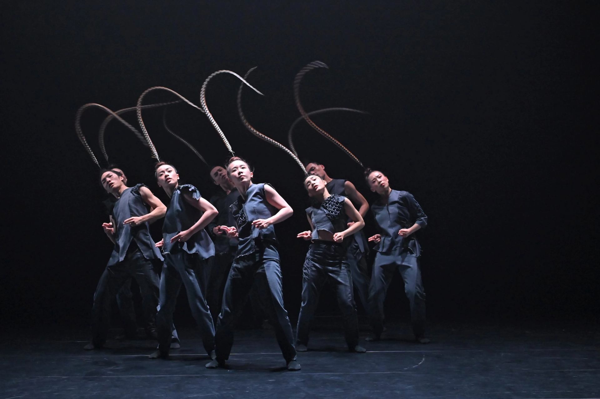 Dancers in dark costumes swing long braids overhead on a dimly lit stage.