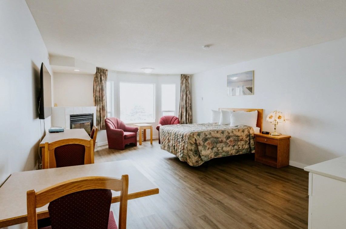 Hotel room with floral bedspread, fireplace, armchairs by large window, and wooden furniture.