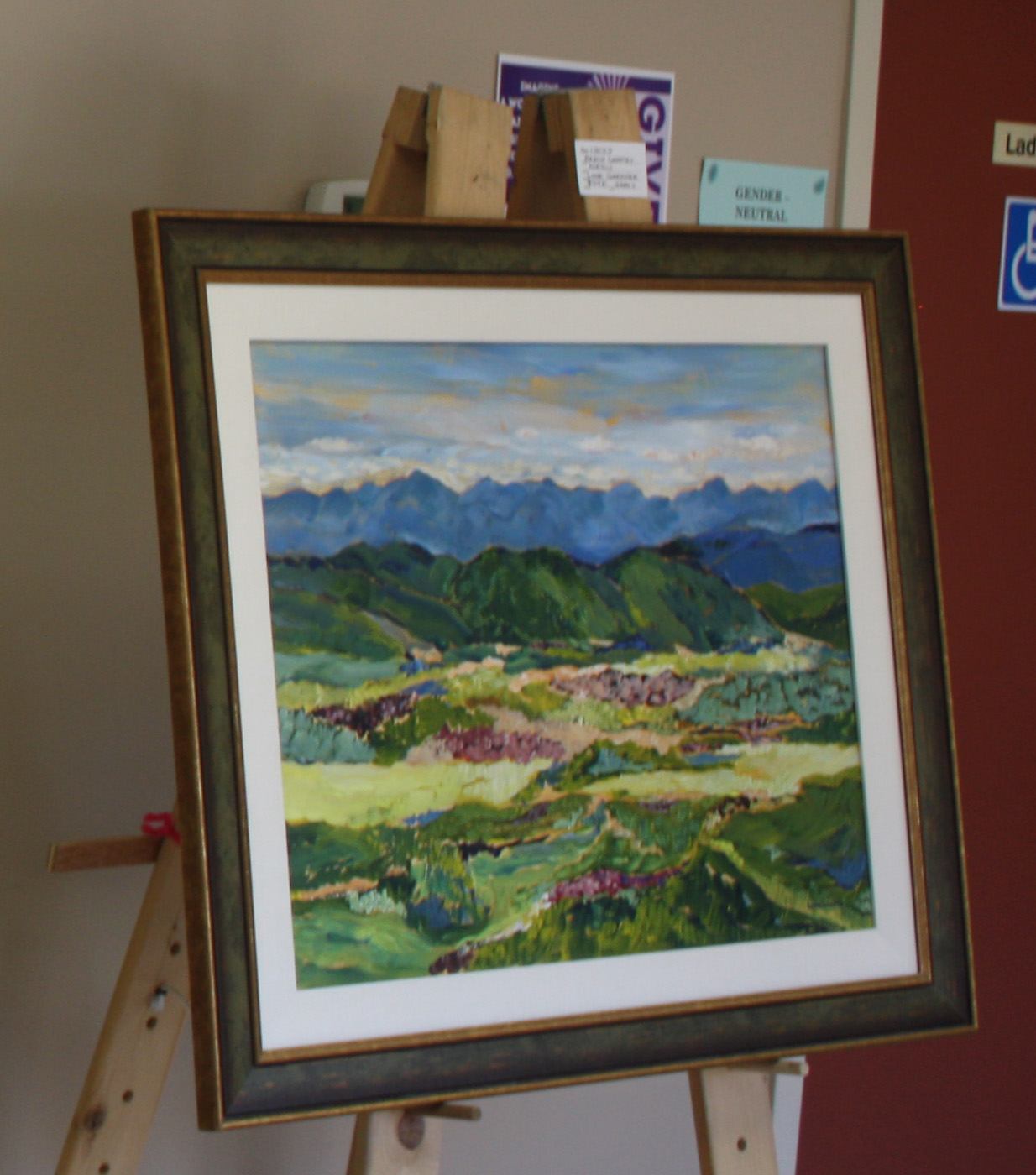 Framed landscape painting of green hills and distant mountains on an easel indoors.