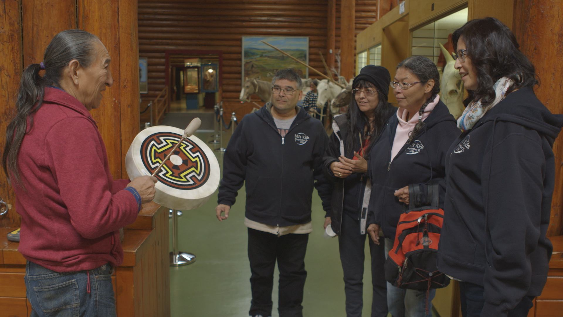 Elderly person holds painted hand drum while four visitors watch inside wooden building.