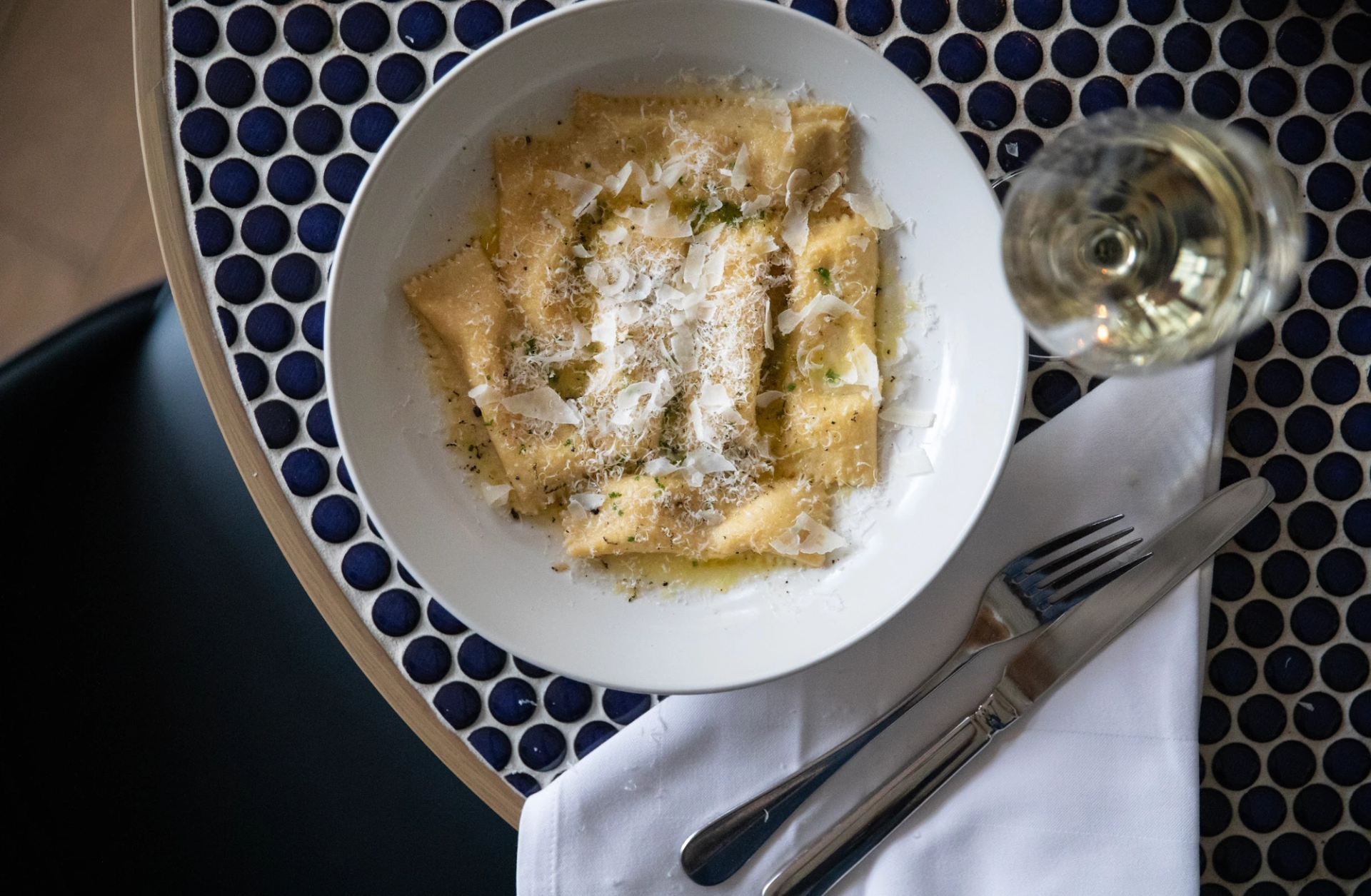 Overhead shot of ravioli topped with cheese and herbs, a glass of white wine, and cutlery on a blue and white patterned table.