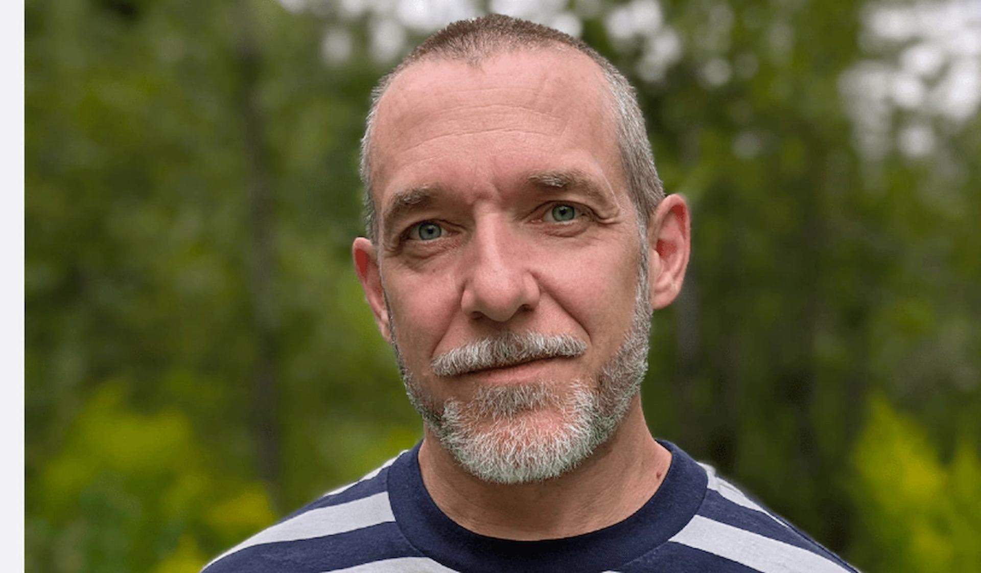 Person with short hair and beard stands outdoors before blurred green trees.