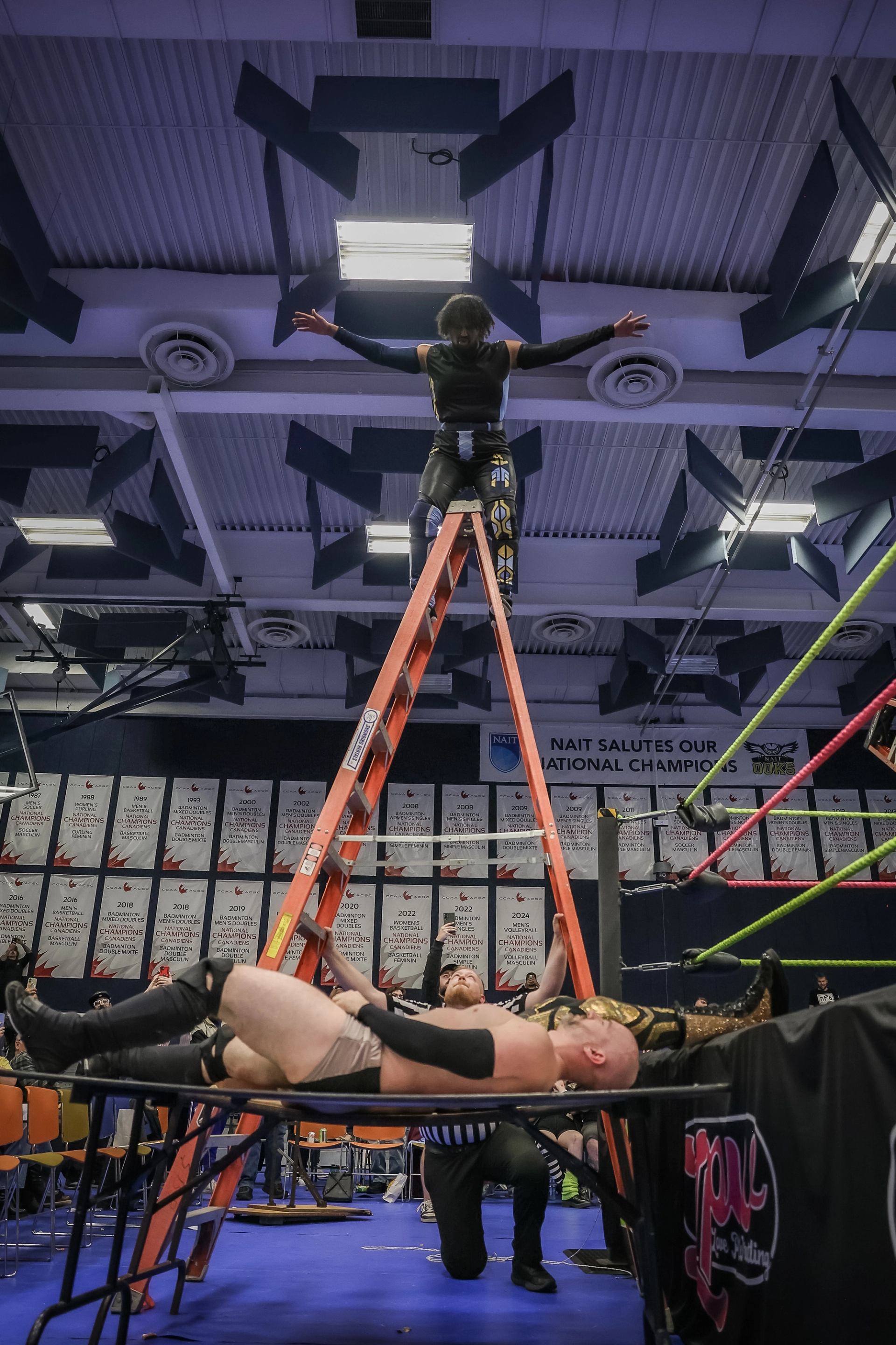 Professional wrestler stands atop tall ladder above opponent lying on broken ringside table.