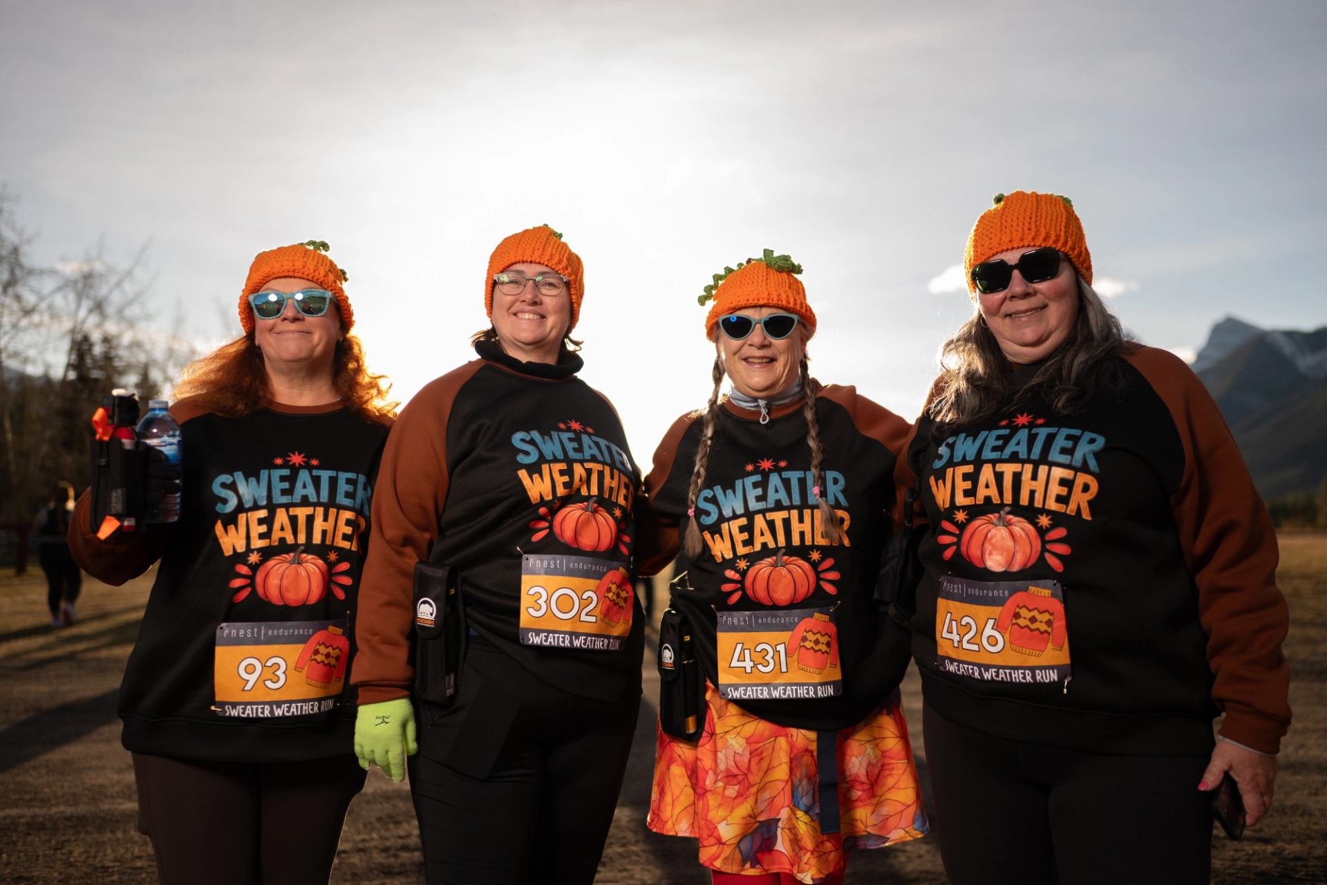 Four participants wearing matching Sweater Weather shirts and pumpkin-themed hats at a trail race.