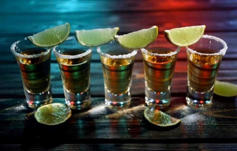 Row of shot glasses with salted rims and lime wedges on wooden table.