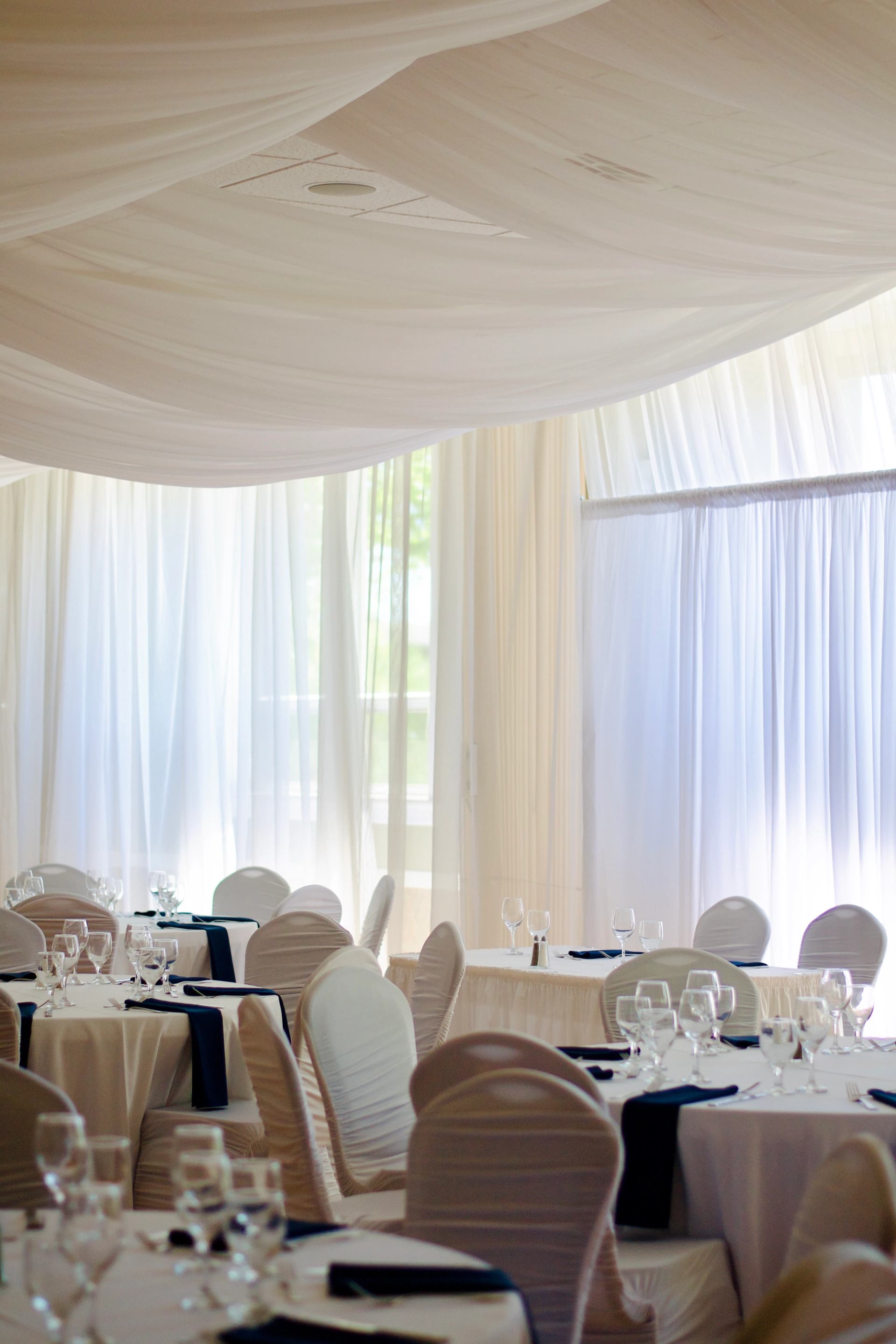 Tables set with glassware and white linens near large windows with sheer curtains