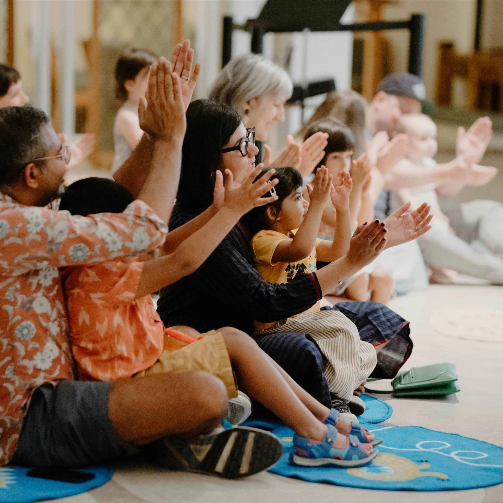 Group seated on mats indoors, clapping hands in a shared activity.