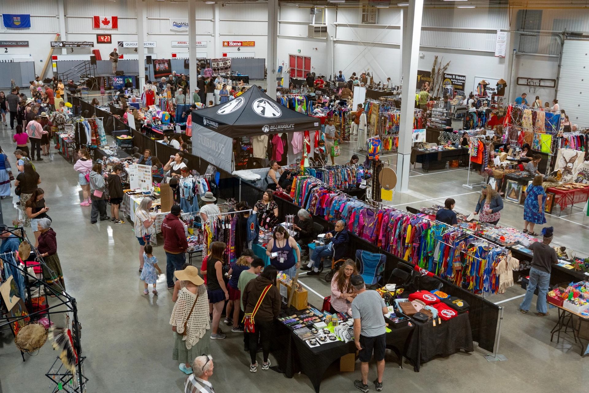 Indoor market with people browsing colourful clothing and craft booths under Canadian flag.