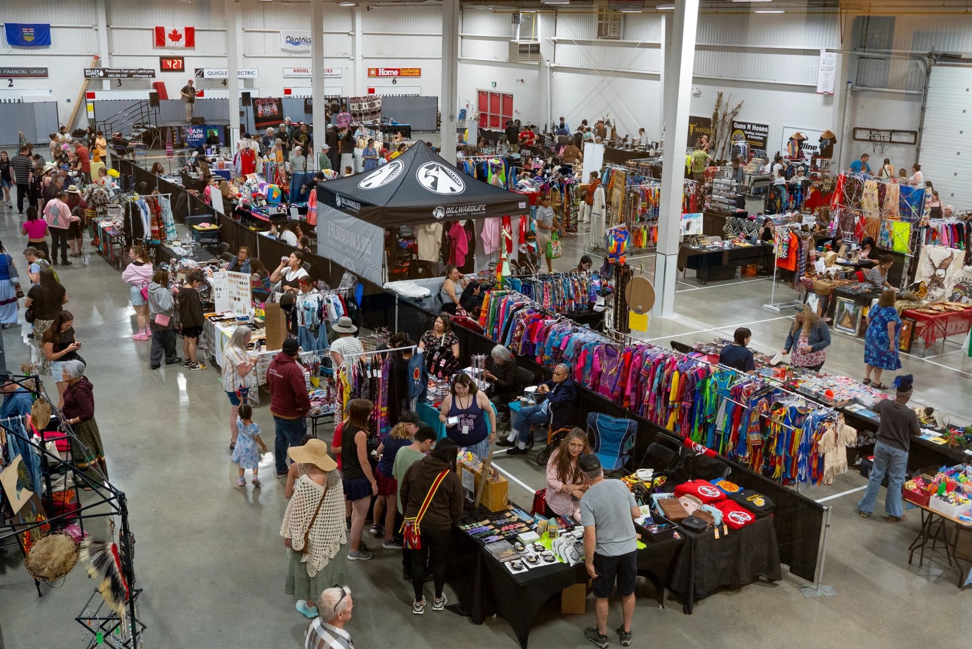 Indoor market with people browsing colourful clothing and craft booths under Canadian flag.