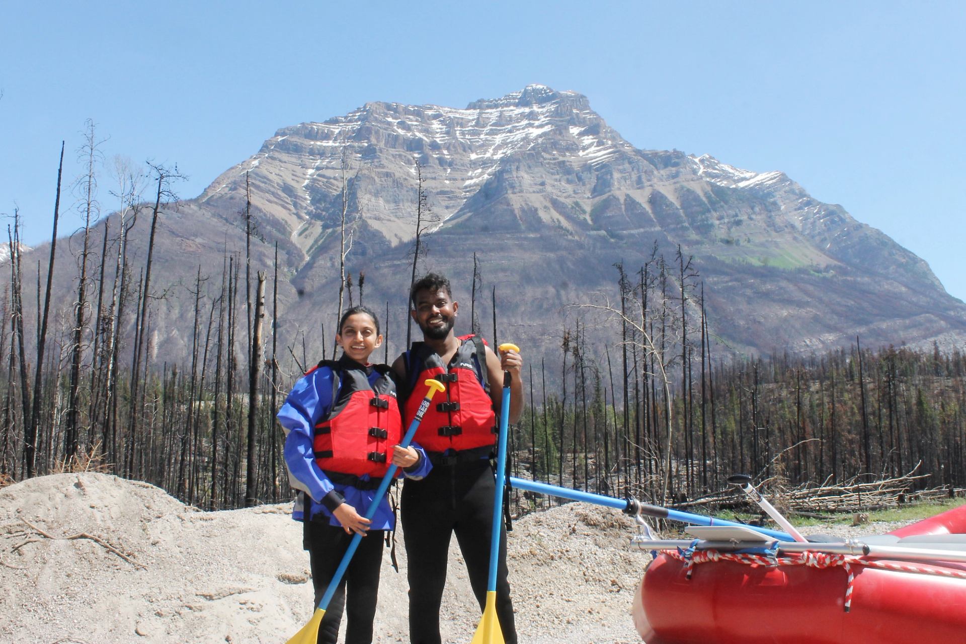 Two people in rafting gear stand beside a red raft with a tall mountain rising behind them.