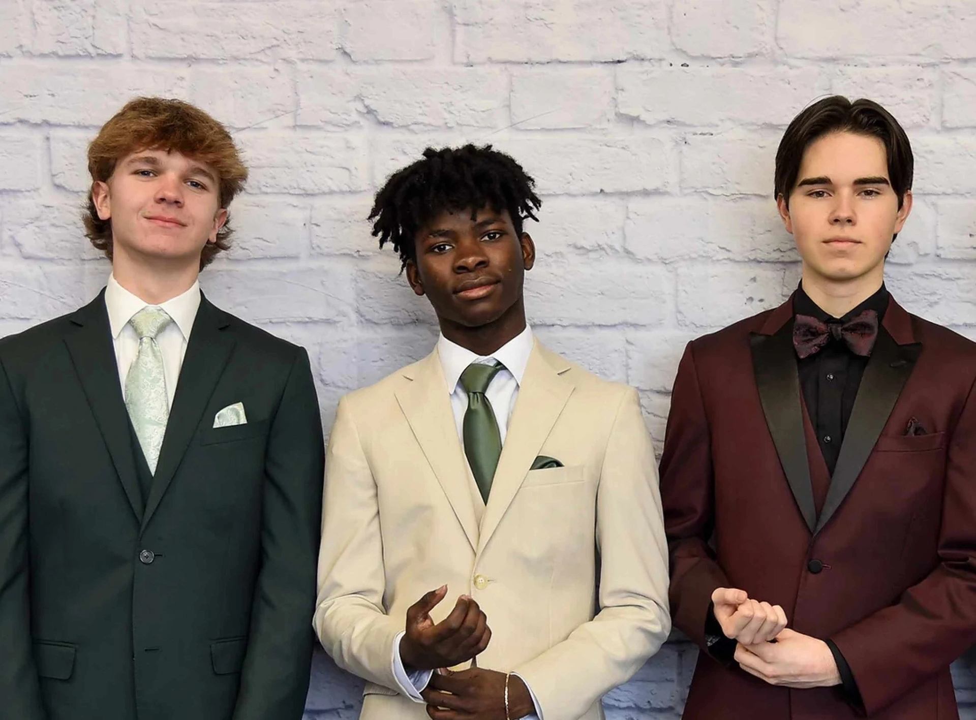 Three people wearing formal suits standing against a light brick wall.
