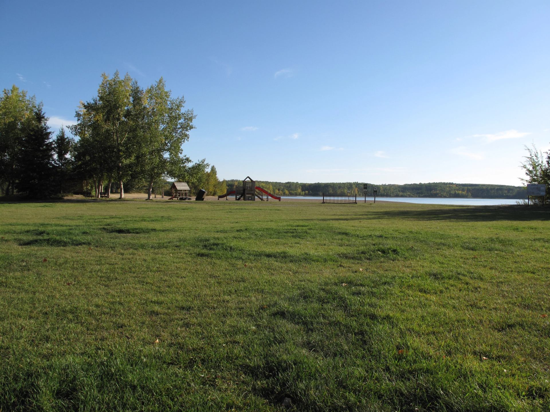 Grassy field with trees, a playground, and a lakeshore under a clear blue sky.