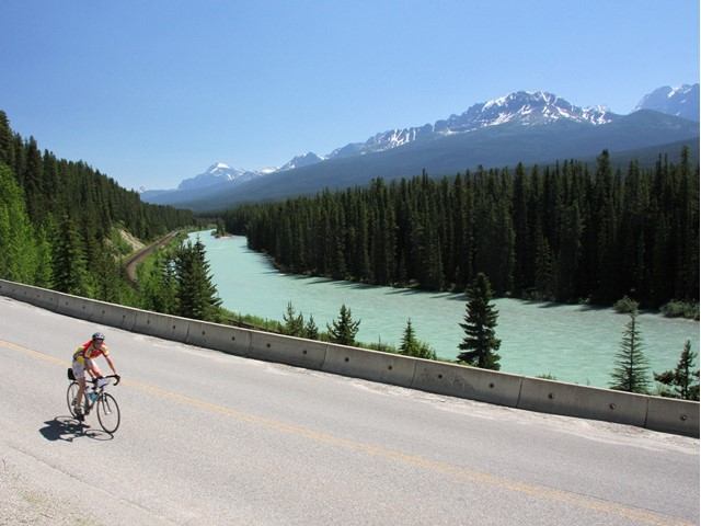Cyclist on riverside road with forest and snow-capped mountains in the background.