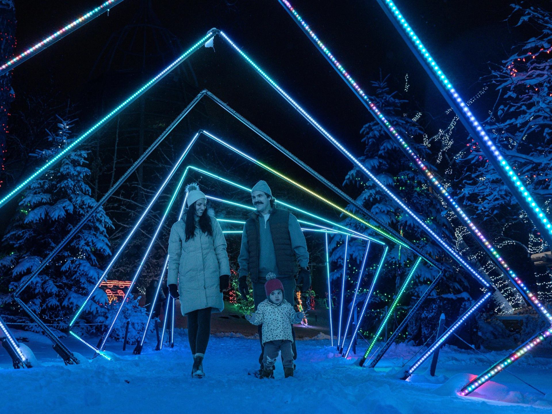 Illuminated triangle light structure with two adults and a child standing outdoors in winter snow.