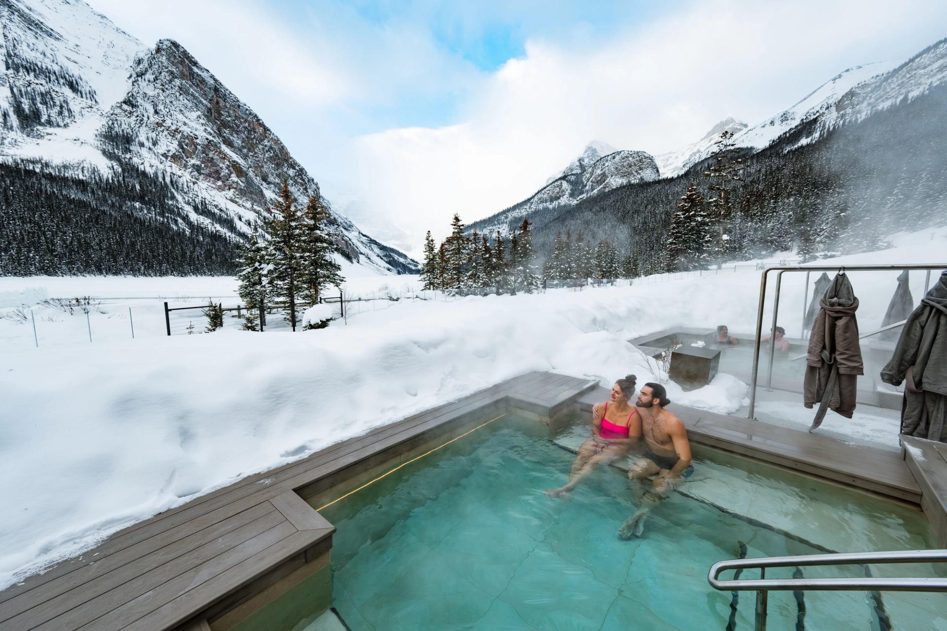 People relax in an outdoor hot pool surrounded by snow-covered mountains and evergreens.