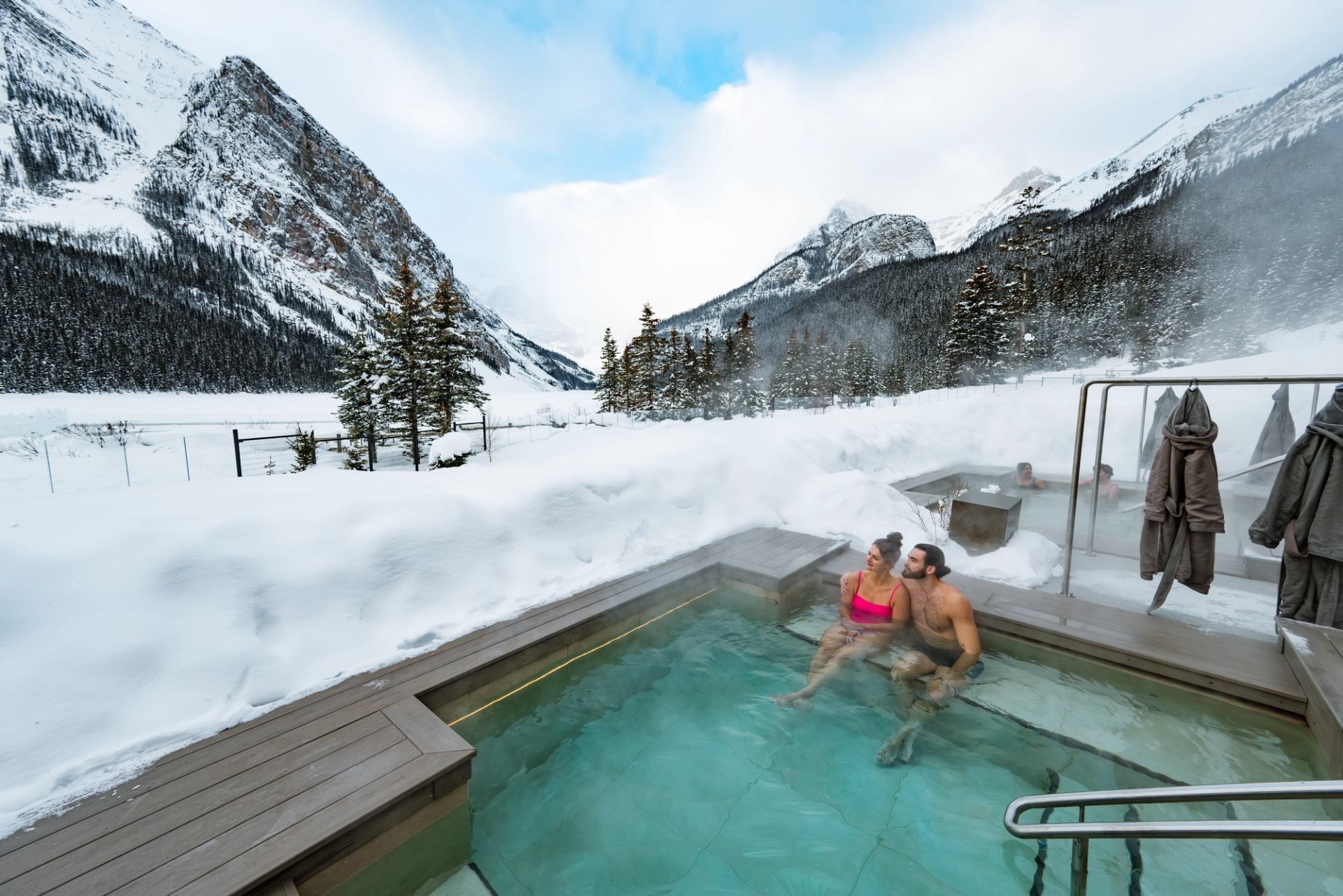 People relax in an outdoor hot pool surrounded by snow-covered mountains and evergreens.