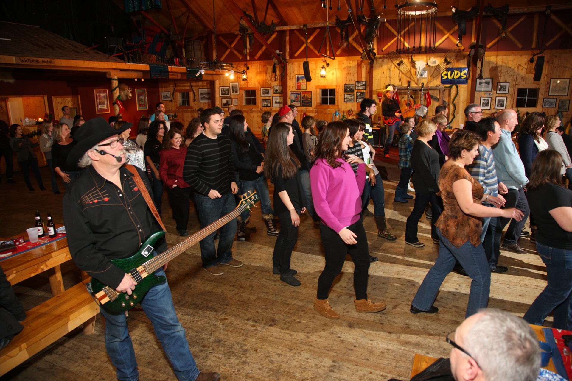 People line dance in rustic hall with live guitar music and western decor.