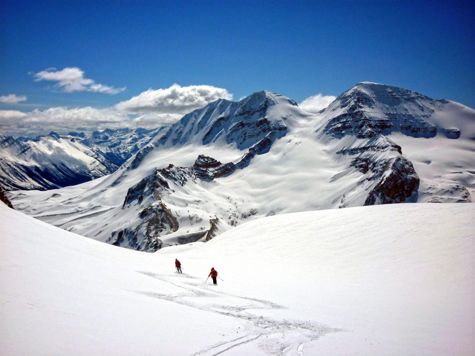Backcountry skiers descend a wide snowy slope below rugged, snow-covered mountain peaks.