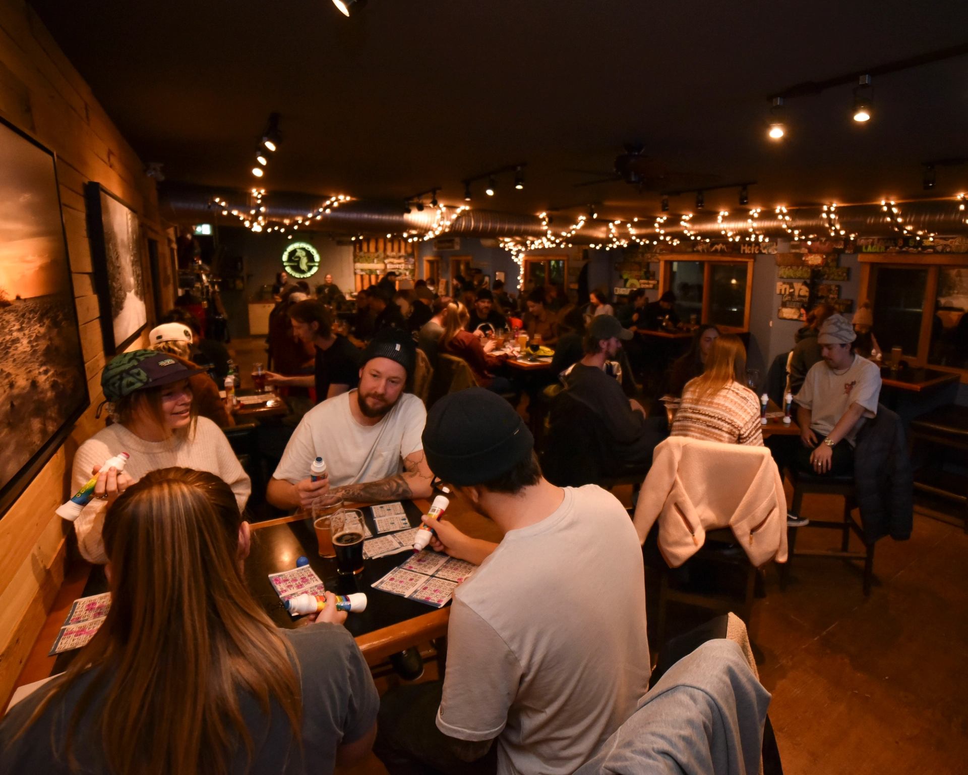 Crowded pub patrons play bingo at tables beneath strings of warm lights.