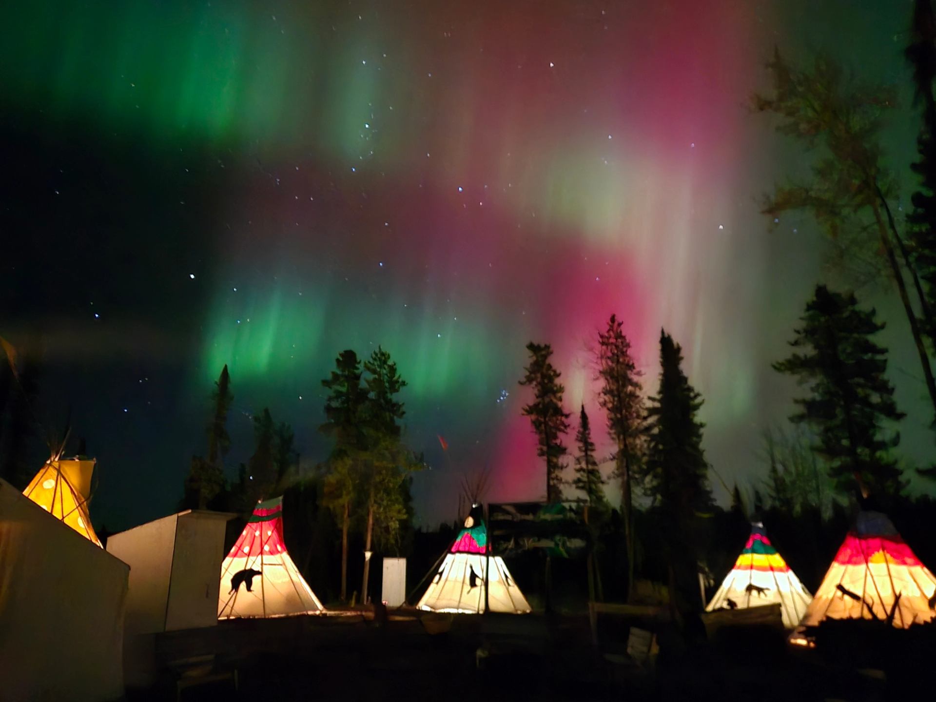 A curtain of Aurora dancing above 5 painted teepees in colors of pink, white, and green
