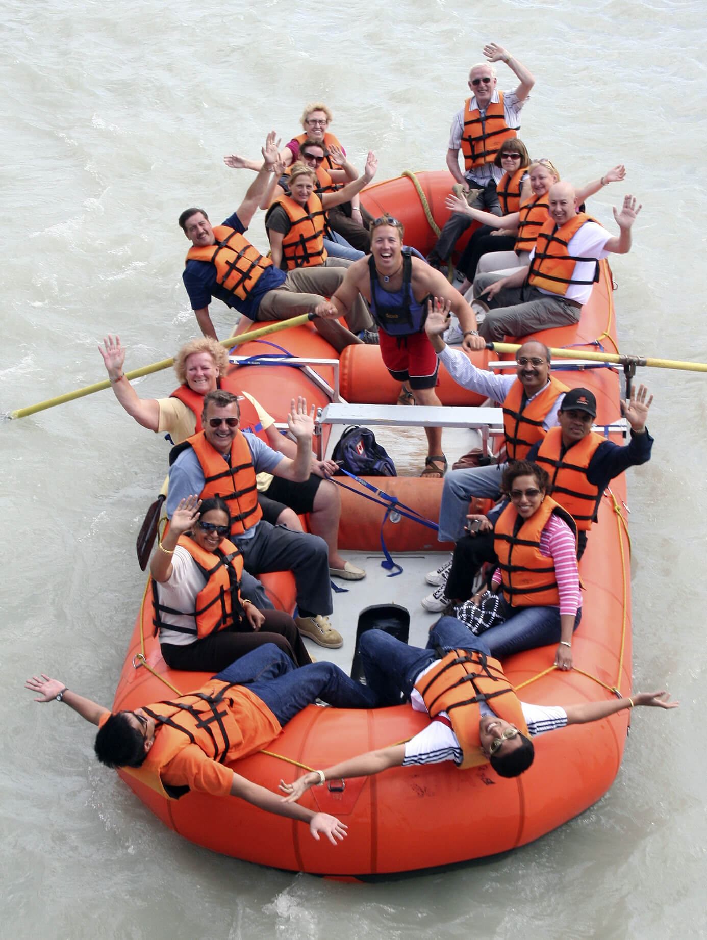 Group of rafters in orange life jackets wave and smile while sitting together on a raft on calm water.