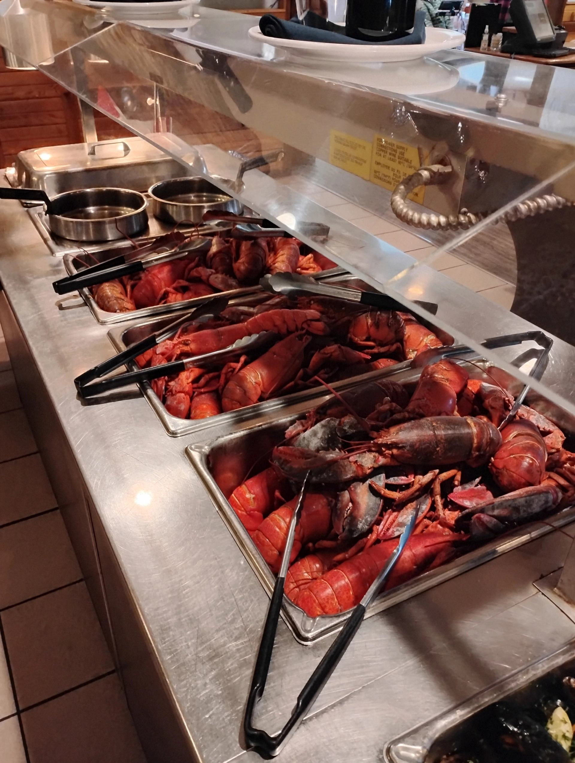 A buffet line with multiple trays of cooked lobsters and serving tongs.