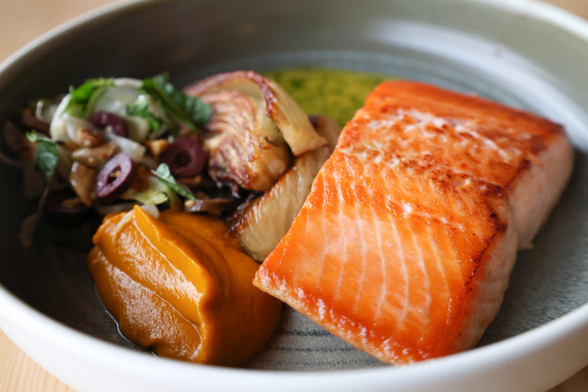 Seared salmon with roasted vegetables and smooth orange puree in a gray bowl.