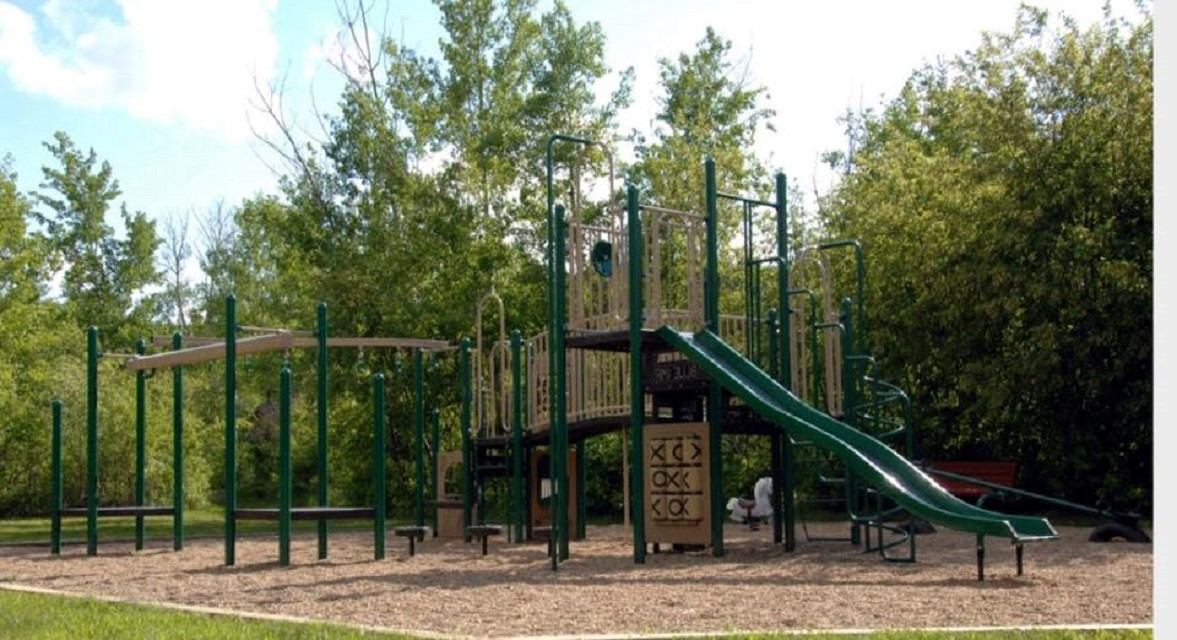 Green playground with slides and climbing bars surrounded by trees and wood chip ground.
