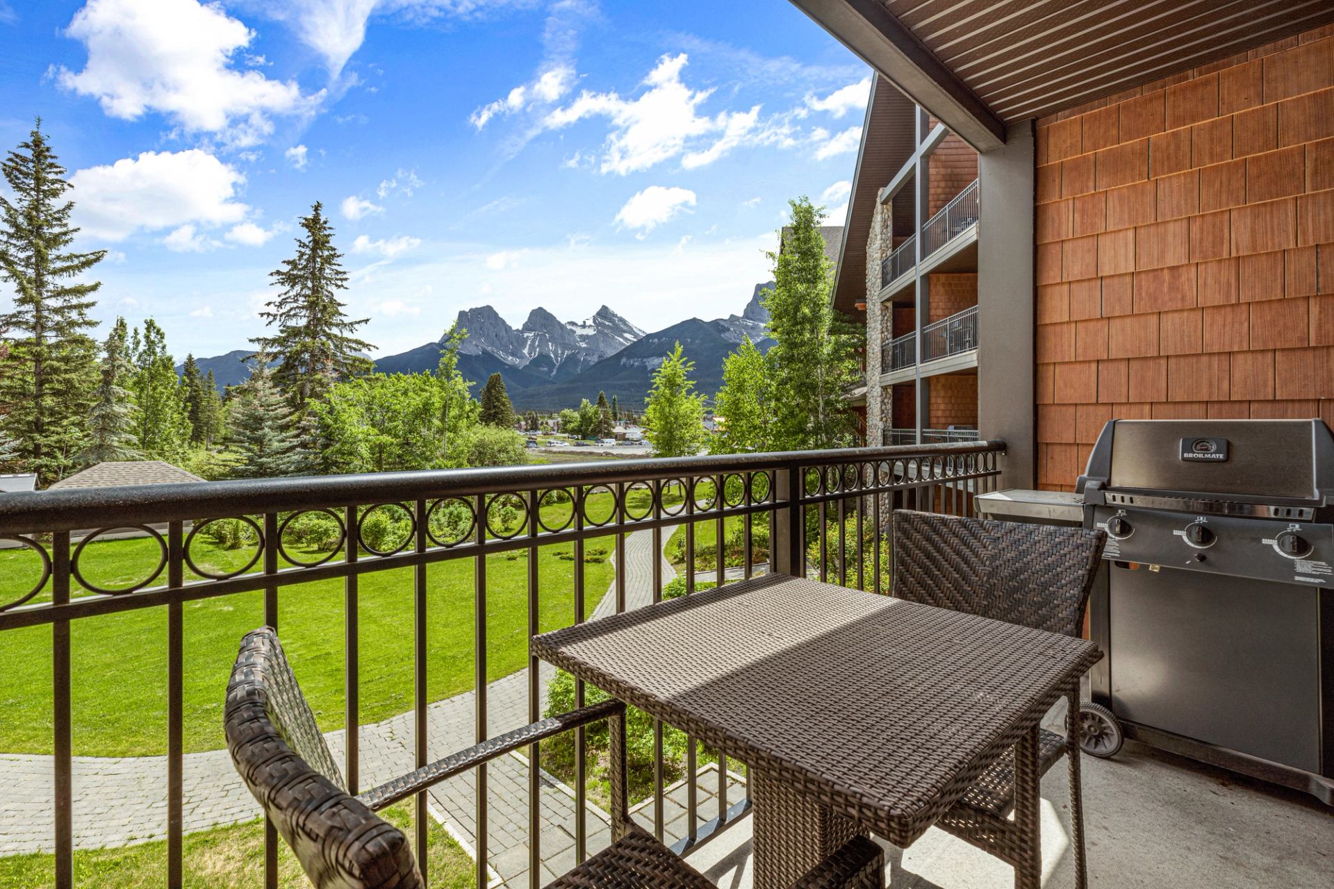 Furnished balcony with barbecue overlooks green grounds, evergreen trees and rocky mountains.