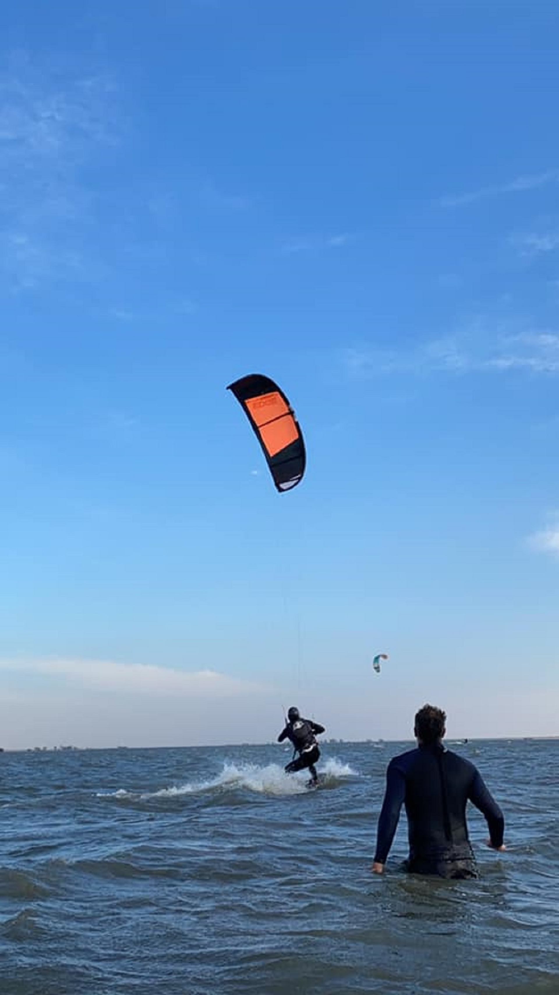 Kiteboarder rides across choppy water while another person stands waist‑deep watching, with kites flying overhead.