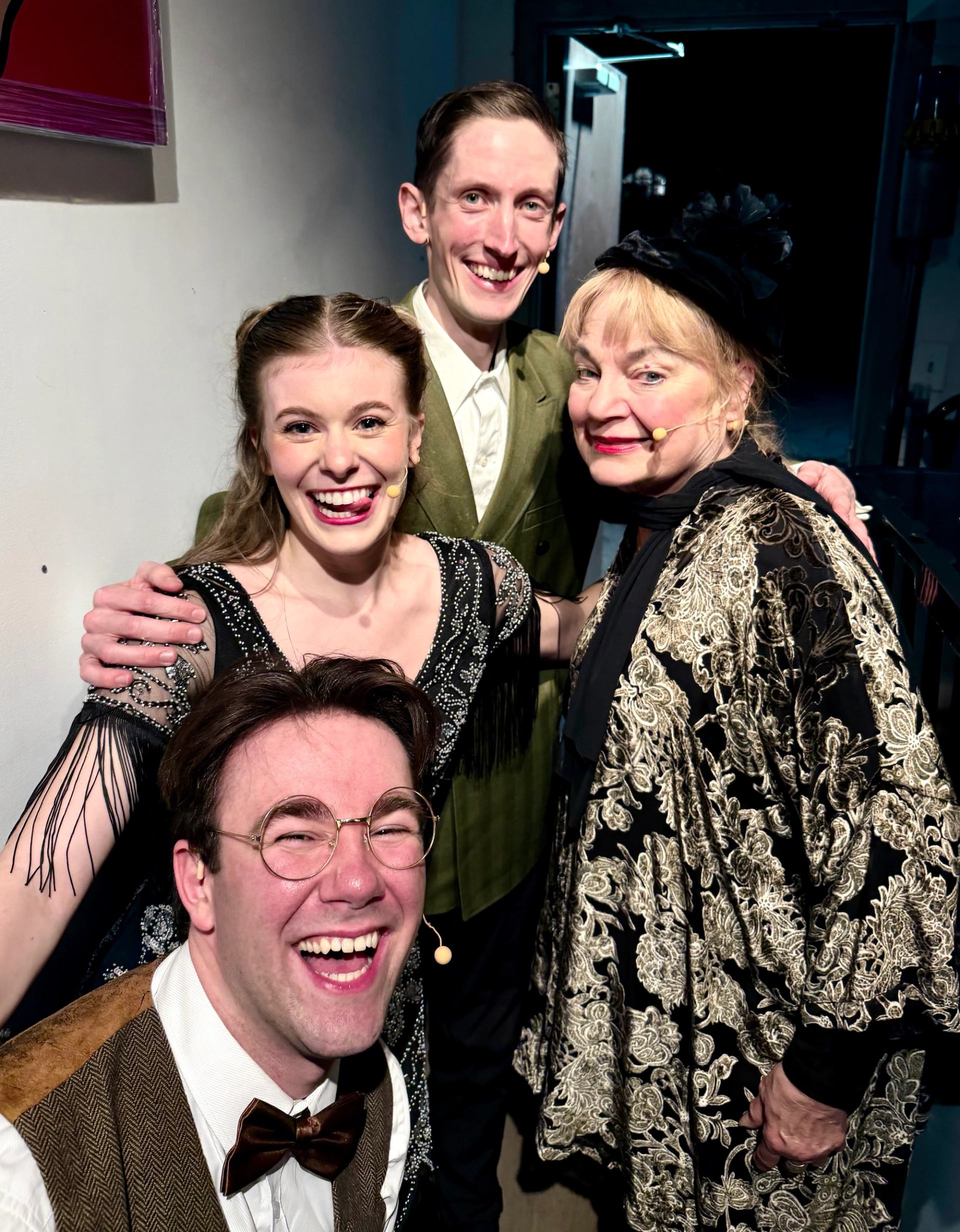 Four performers wearing headset microphones pose together in theatrical costumes.