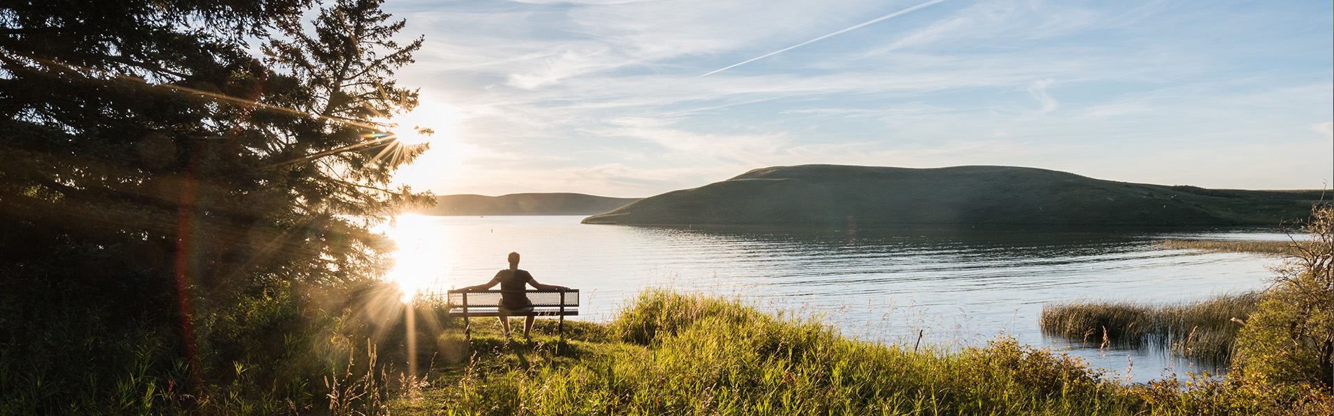 A person sits on a bench overlooking calm lake waters and rolling hills at Elkwater Lake Marina.