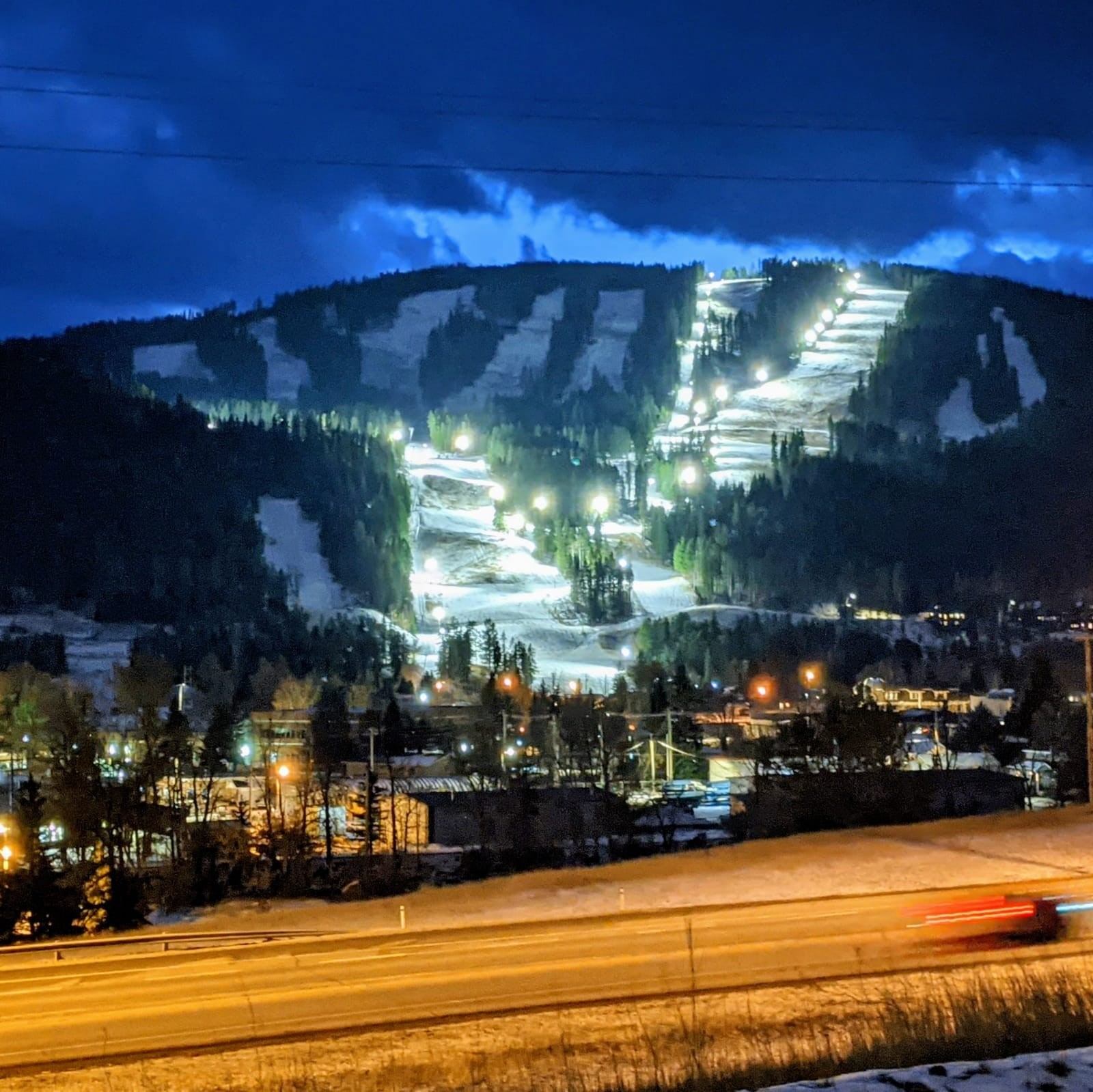 Pass Powderkeg ski hill illuminated at night with glowing trails and surrounding town lights.