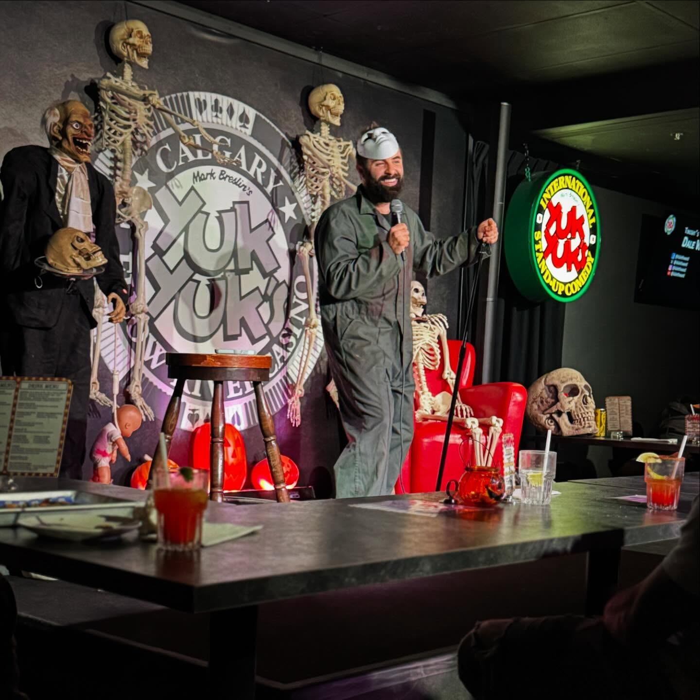 Comedy club stage with performer holding microphone, skeleton decorations, and Yuk Yuk’s logo sign.