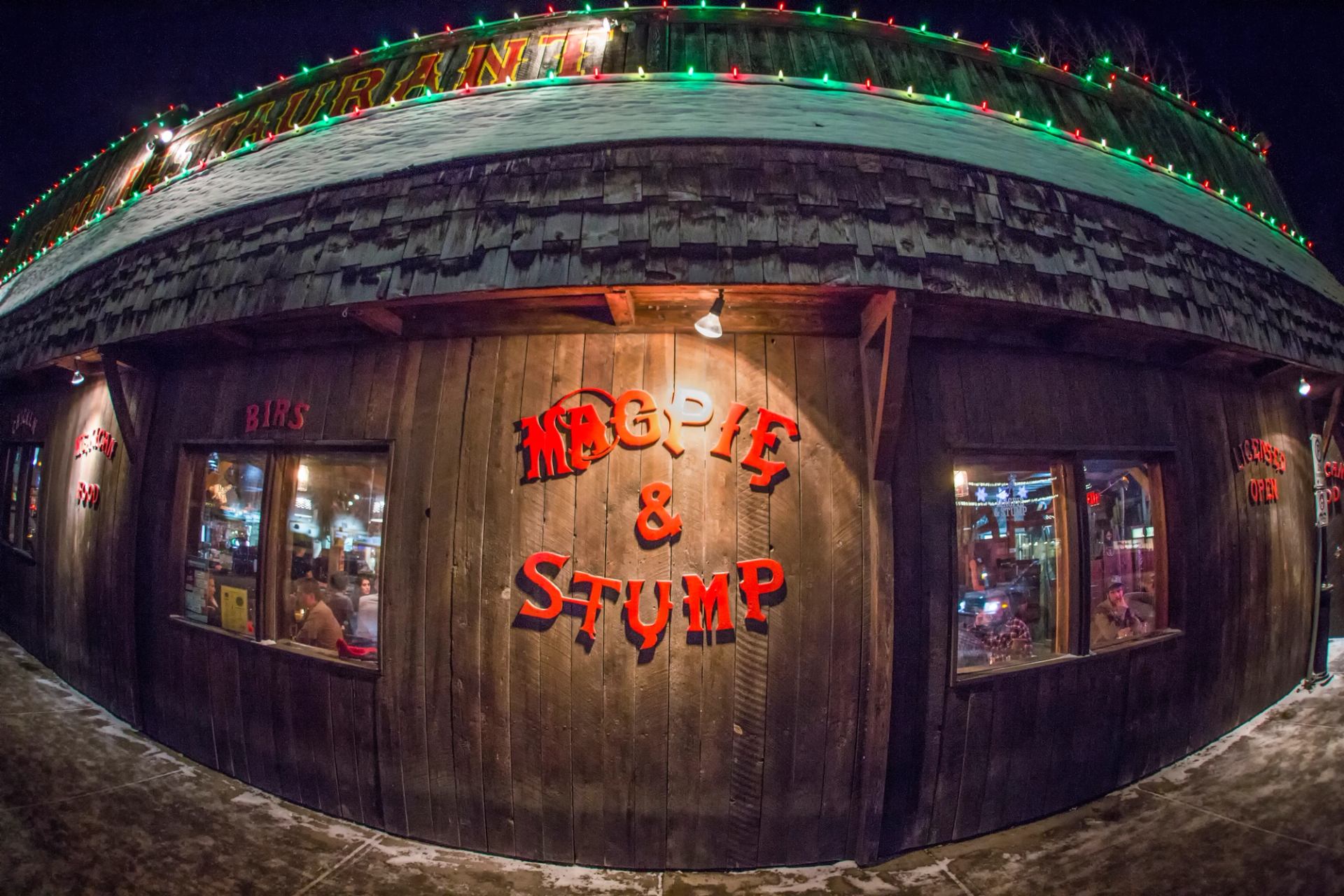 Exterior of the rustic wooden "MAGPIE & STUMP" restaurant at night, with snow on its roof and colorful lights.
