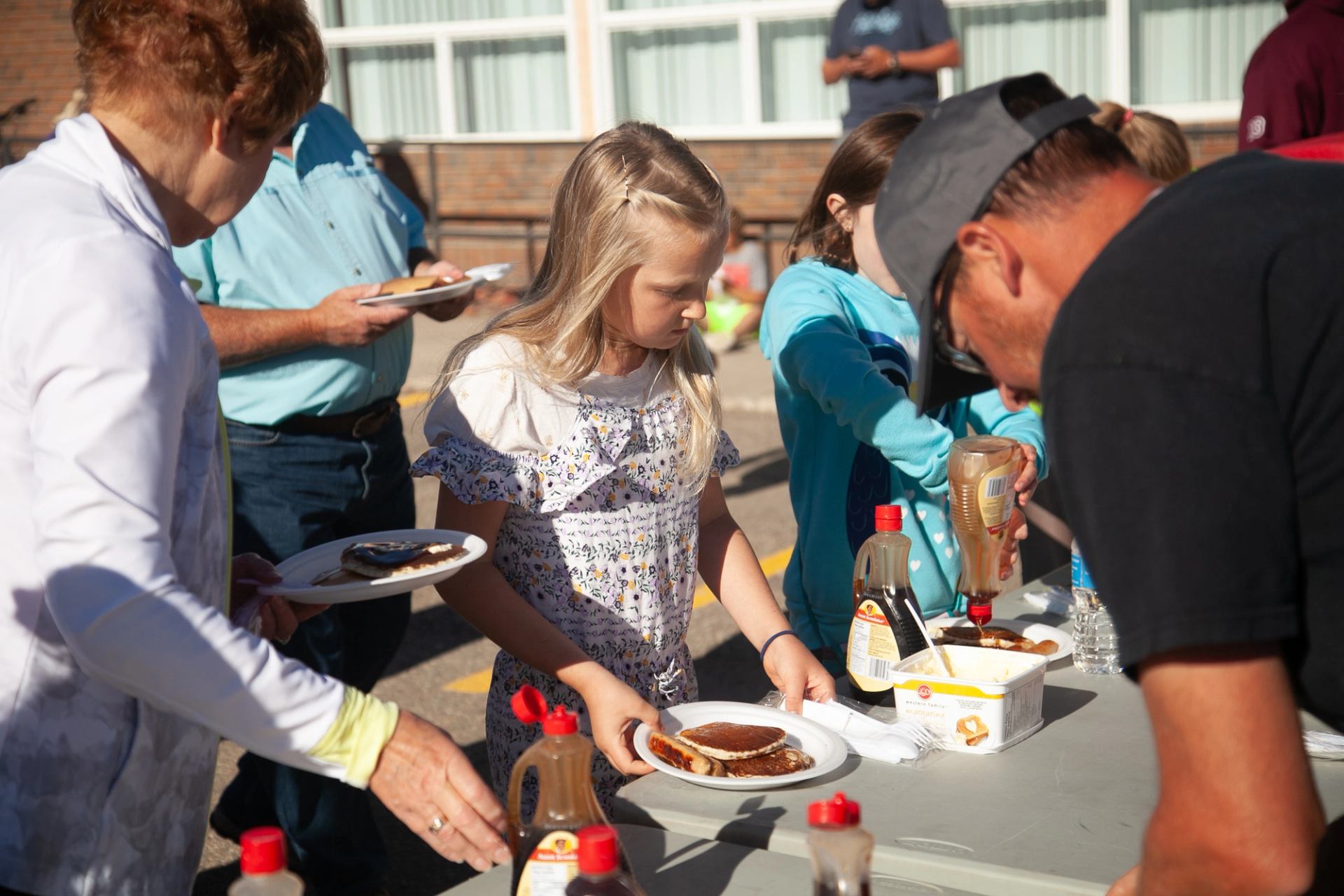 People serve pancakes with syrup at an outdoor table during a community event.